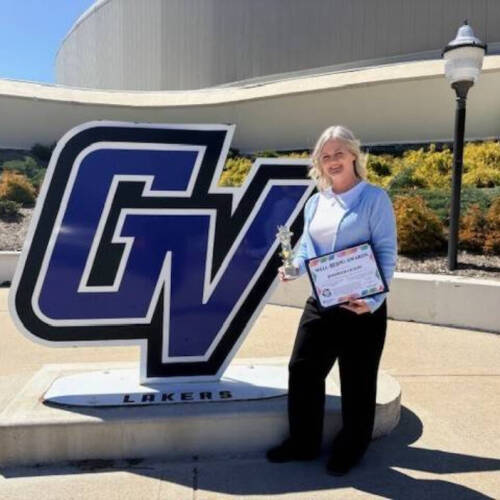 Jen Olson standing in front of the GVSU Sign holding her Golden Apple Award